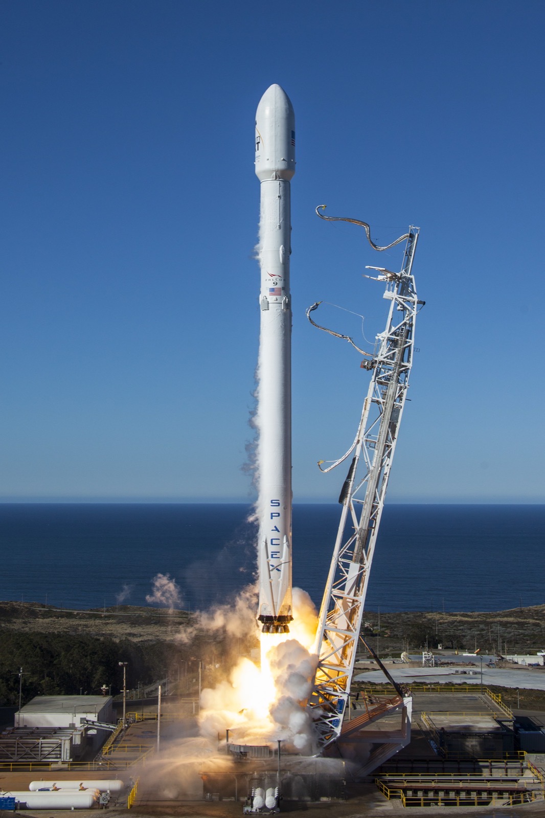 A white SpaceX Falcon 9 rocket lifting off from a coastal launch pad in daylight, with the Pacific Ocean visible behind the strongback tower