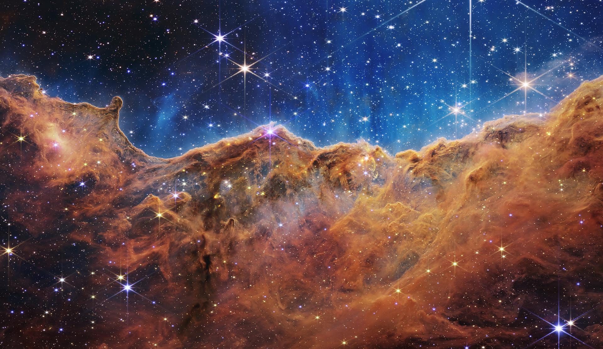 JWST's 'Cosmic Cliffs' image of the Carina Nebula's NGC 3324 star-forming region, showing towering mountains of blue-orange gas and dust backlit by ultraviolet radiation from massive newborn stars, with hundreds of previously invisible protostars emerging from the nebular haze