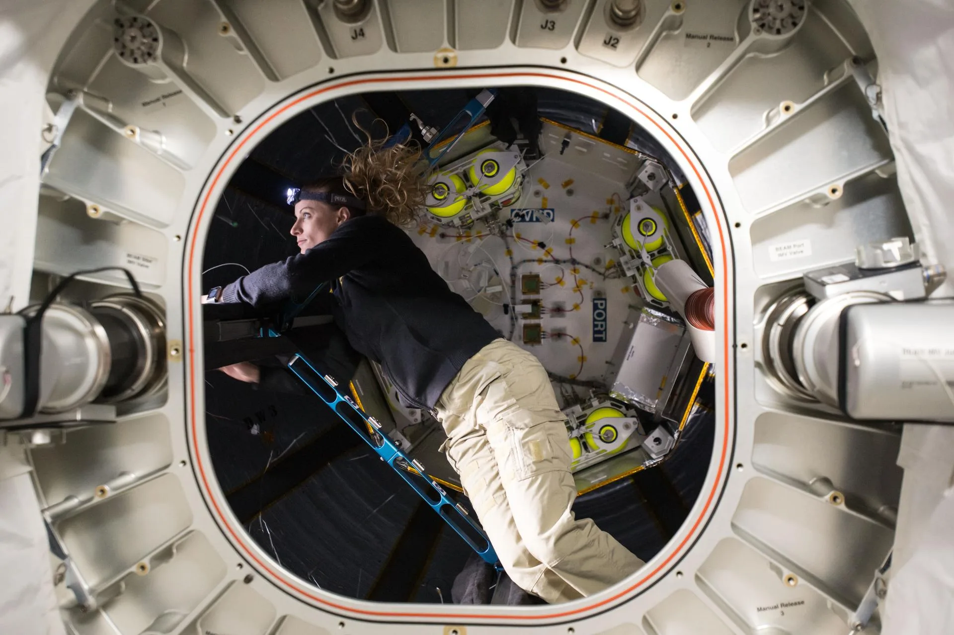 An astronaut wearing a headlamp floats head-first through a rigid metal hatch ring into the dark fabric-walled interior of an expandable module fitted with yellow-capped equipment
