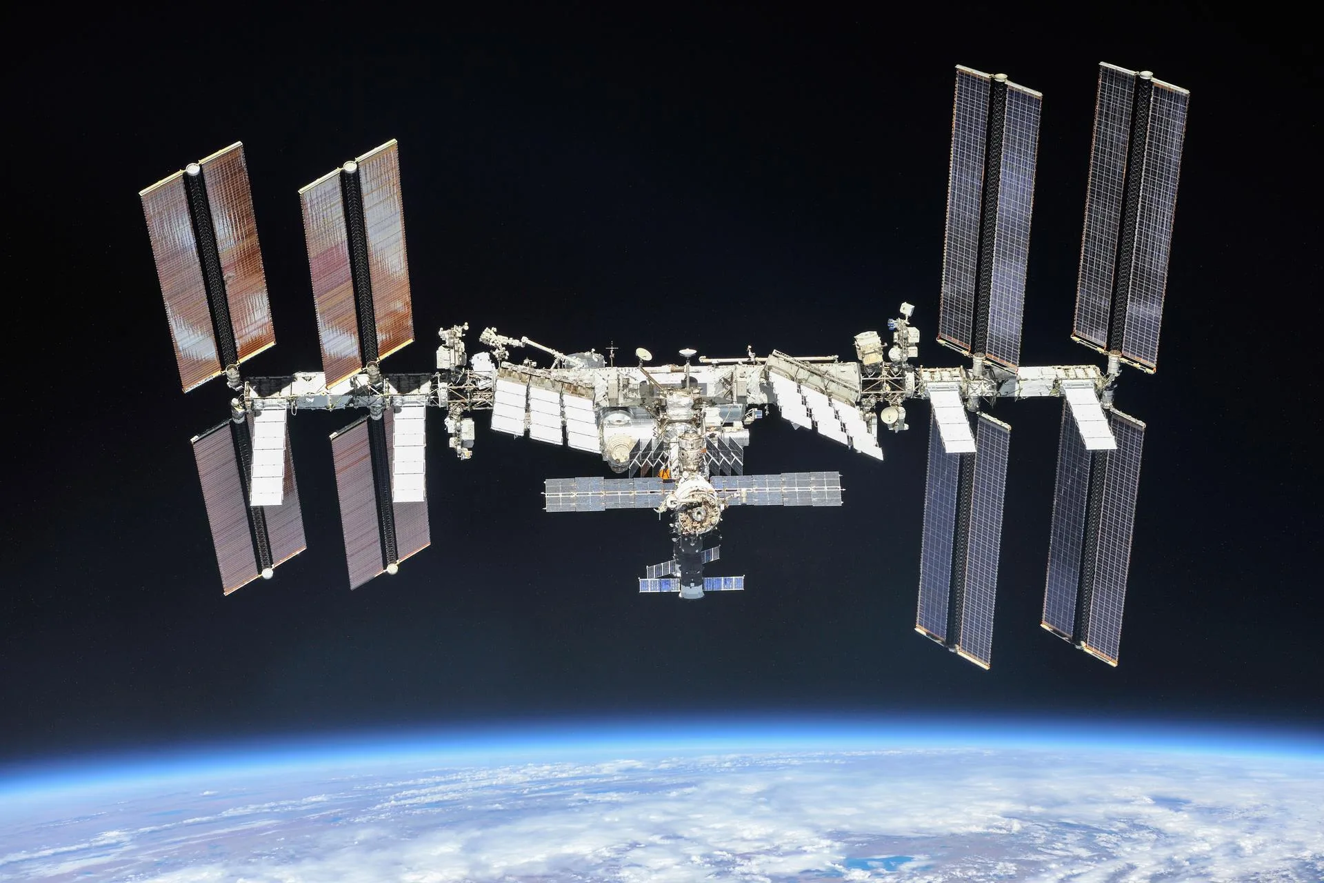 The International Space Station photographed against black space above Earth's blue limb, its four pairs of solar arrays extending from a long central truss with pressurised modules clustered at the centre