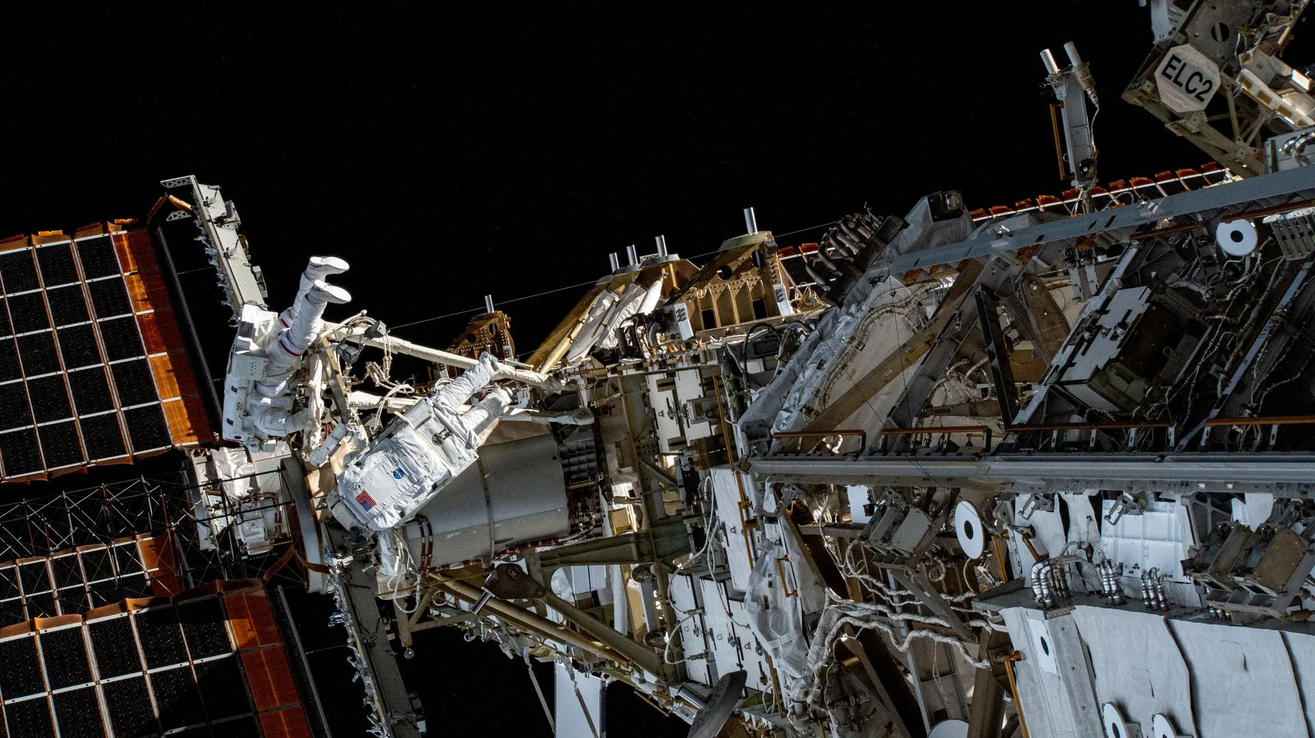 Two spacewalking astronauts in white spacesuits working on the International Space Station's metallic truss structure, with a gold-toned solar array wing extending to the left against black space