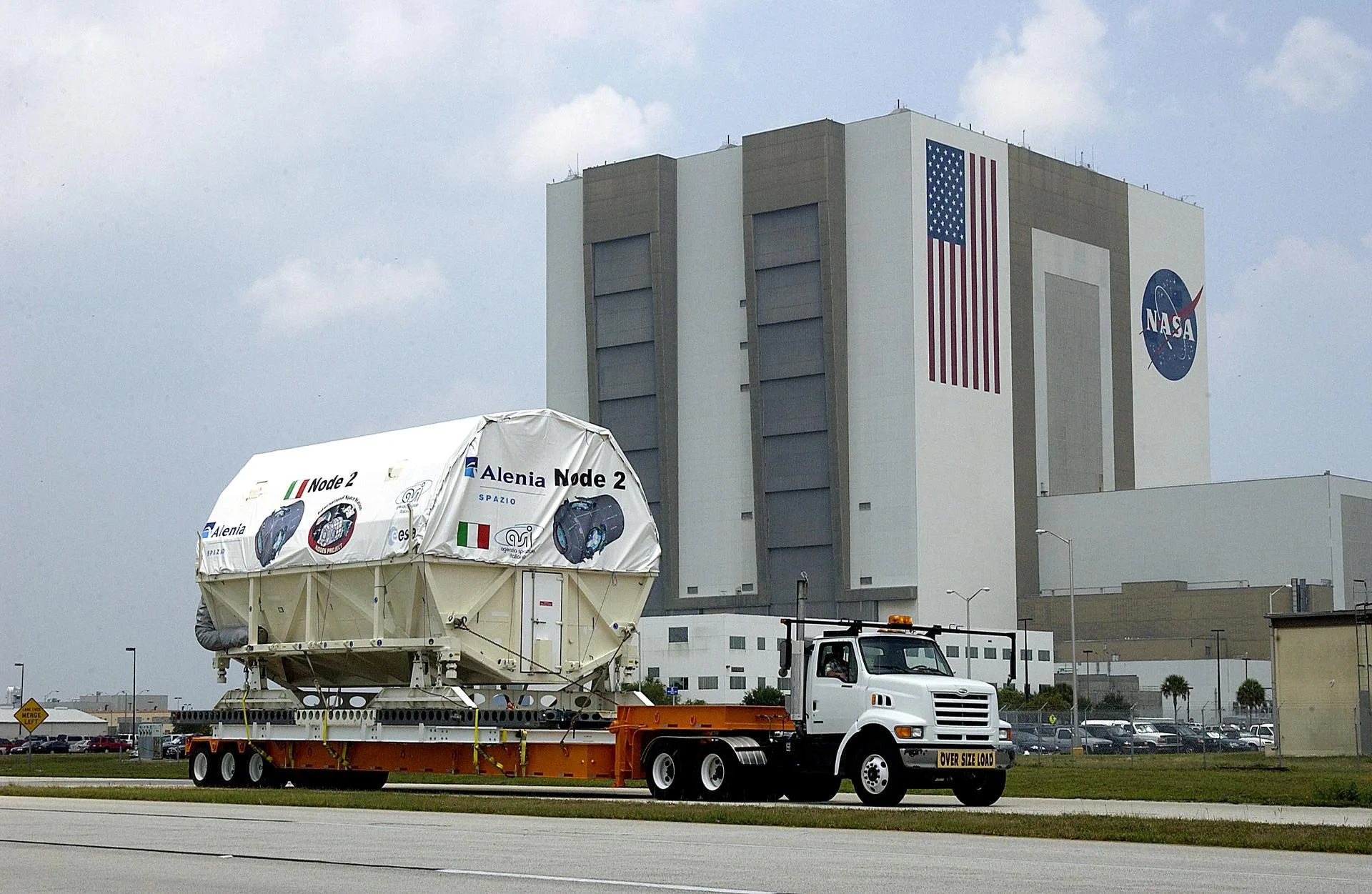 A large white shrink-wrapped cylindrical space station module labelled 'Alenia Node 2' strapped to an orange flatbed truck trailer, driving past NASA's Vehicle Assembly Building