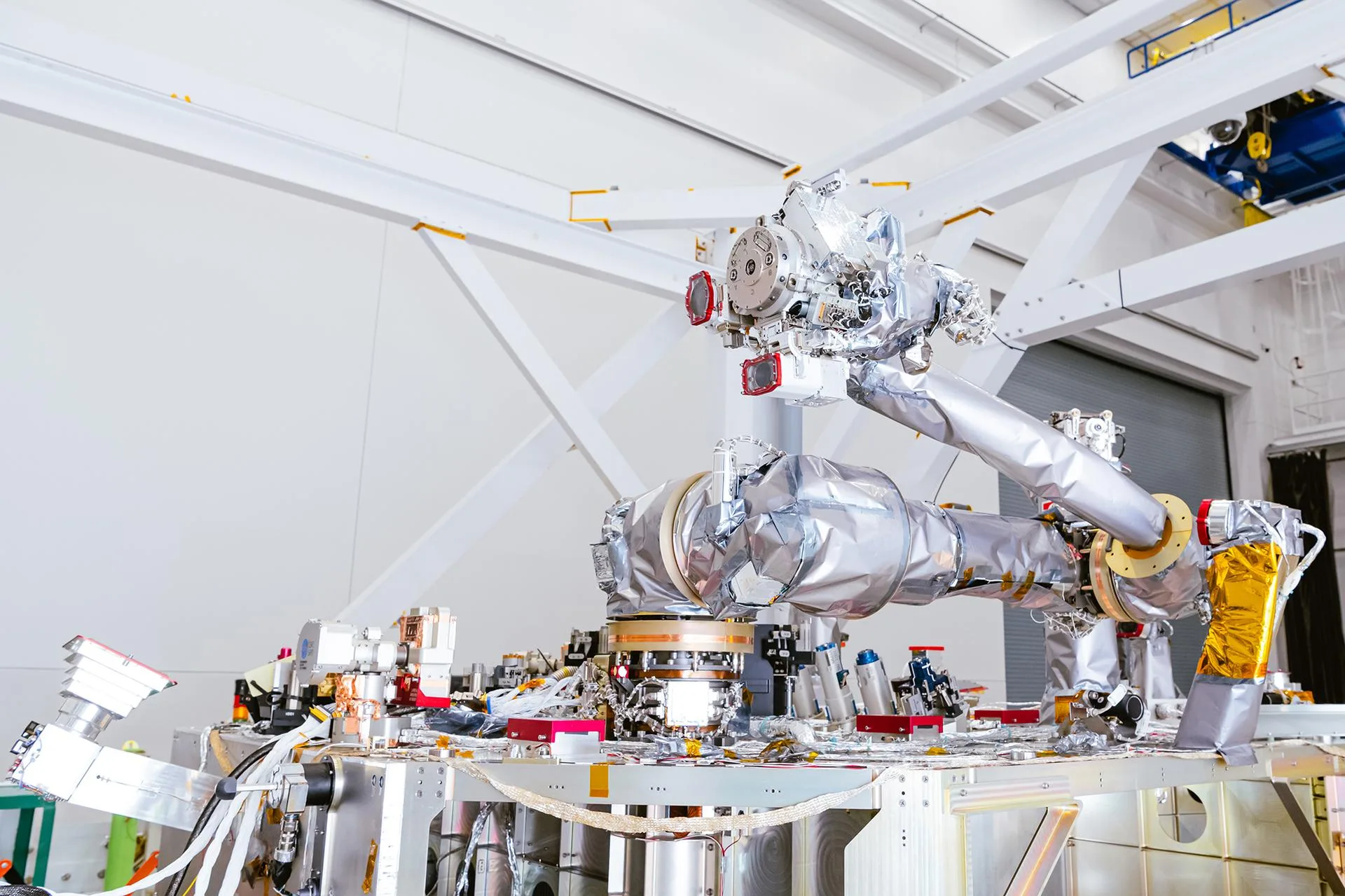 A large silver multi-jointed robotic arm mounted on a densely packed spacecraft deck inside a white cleanroom