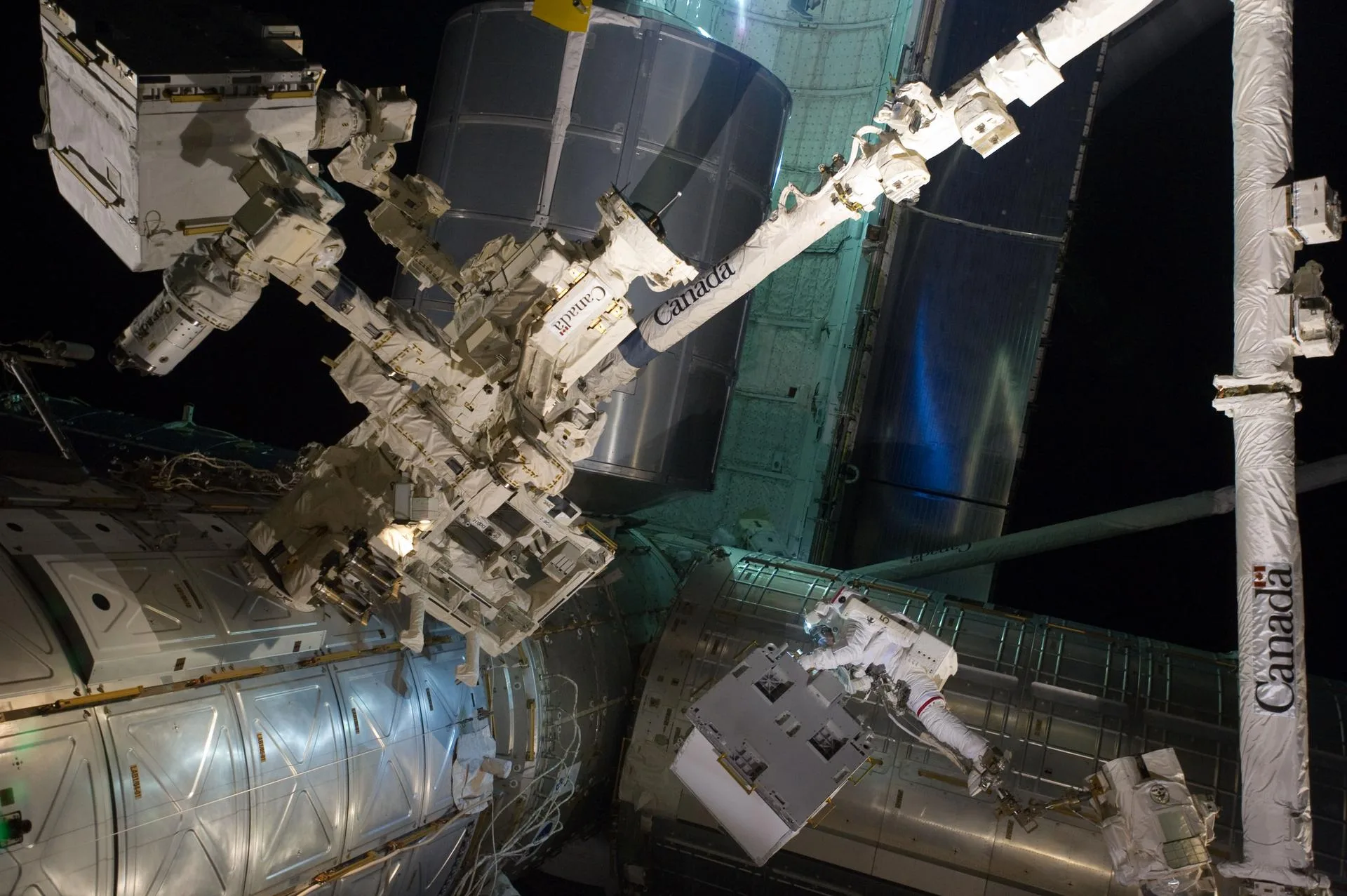 An astronaut on the end of the International Space Station's Canadarm2 holds a white box-shaped payload, with the Dextre robotic manipulator and station modules filling the frame
