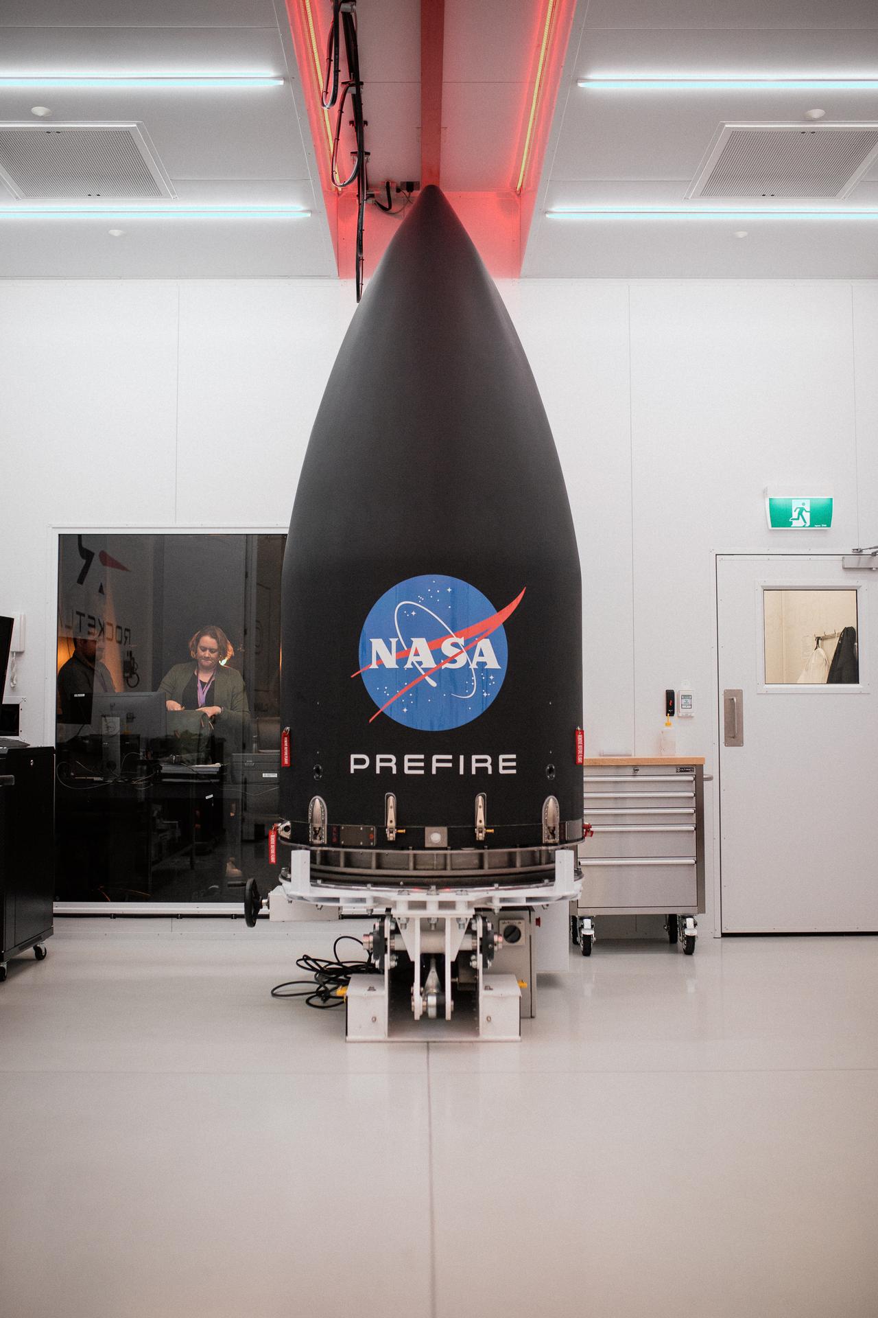A black Electron payload fairing marked with the NASA logo and 'PREFIRE' standing on a transport cradle inside a clean white Rocket Lab integration room, an engineer at a console in the background