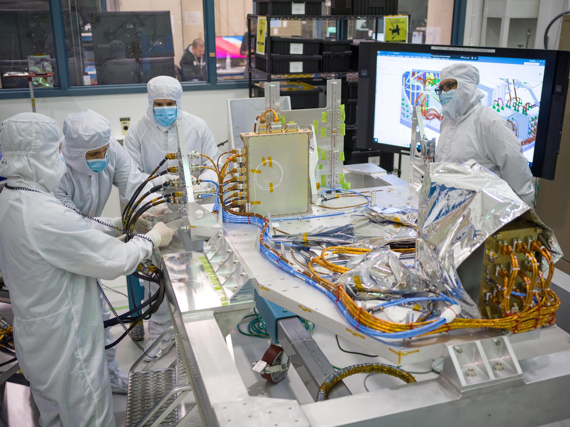 Four engineers in full white cleanroom suits and face shields working at a table assembling Roman's Coronagraph Instrument electronics at NASA's Jet Propulsion Laboratory in December 2022, with yellow anti-static bags and electronic components visible on the table
