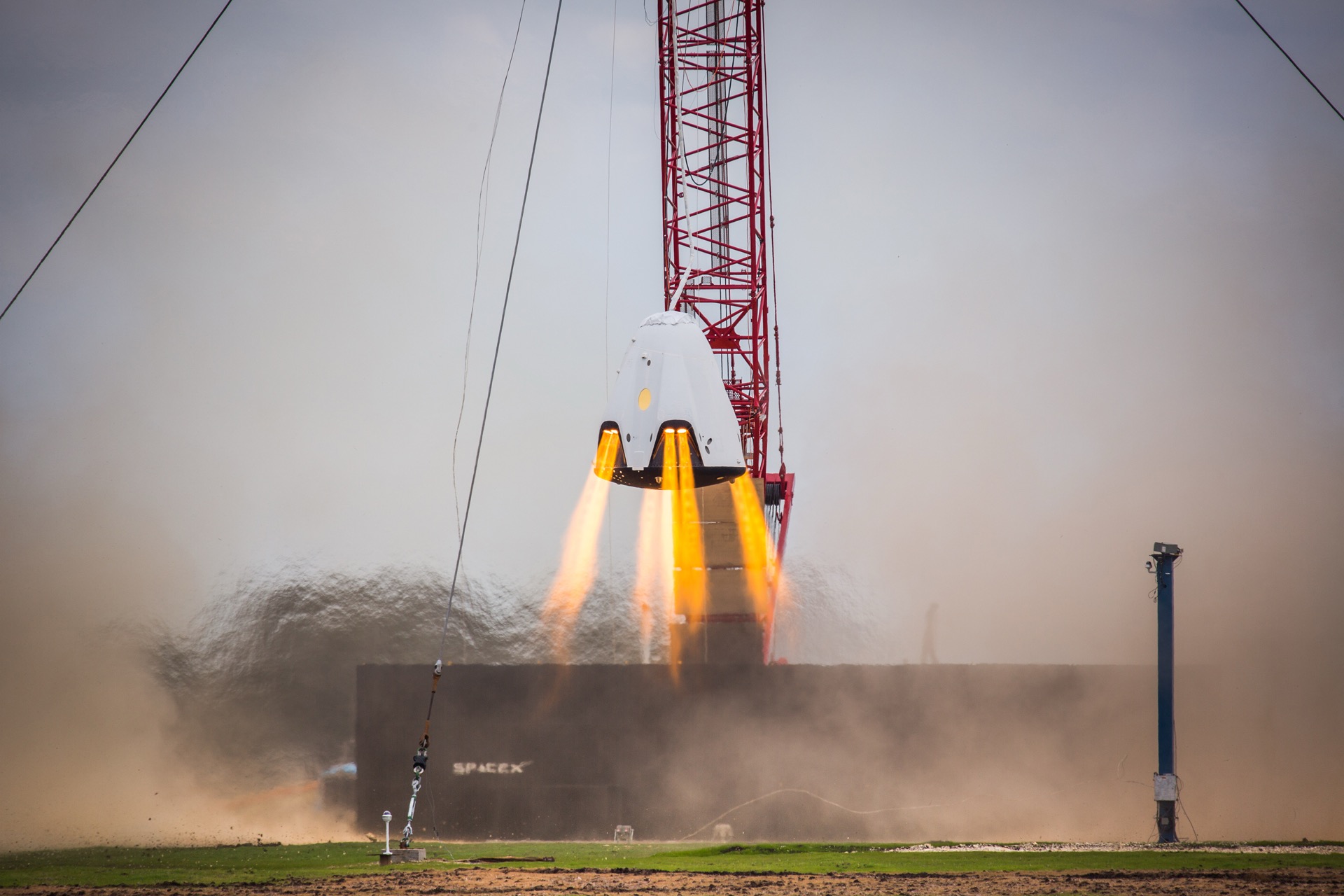 A SpaceX Crew Dragon capsule fires its SuperDraco thrusters in a bright orange plume during a propulsive hover test above a SpaceX test pad