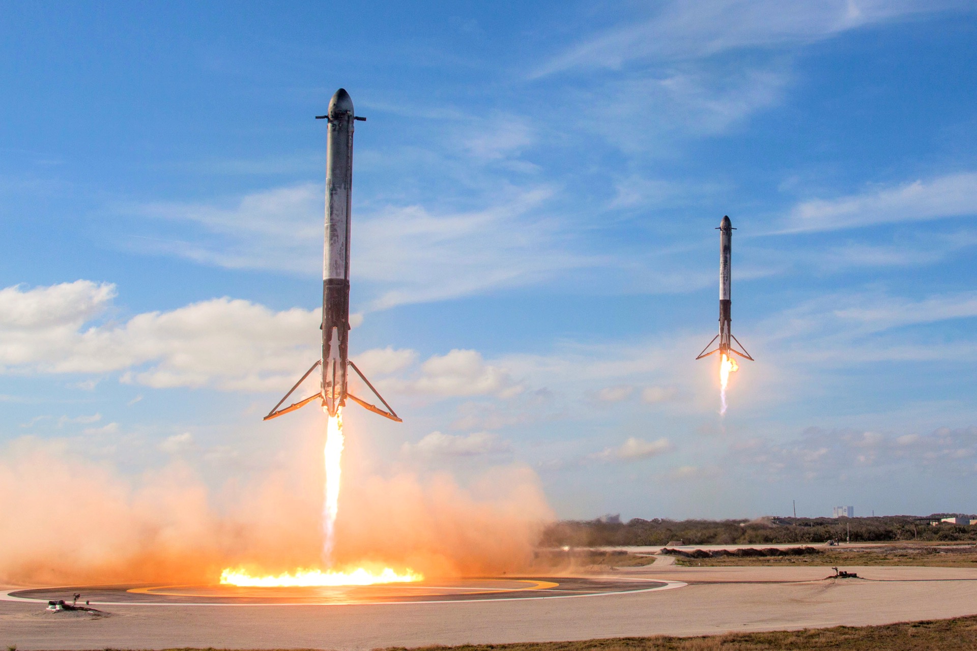 Two SpaceX Falcon Heavy side boosters descend in unison under rocket power toward the landing zones, landing legs deployed, against a blue sky