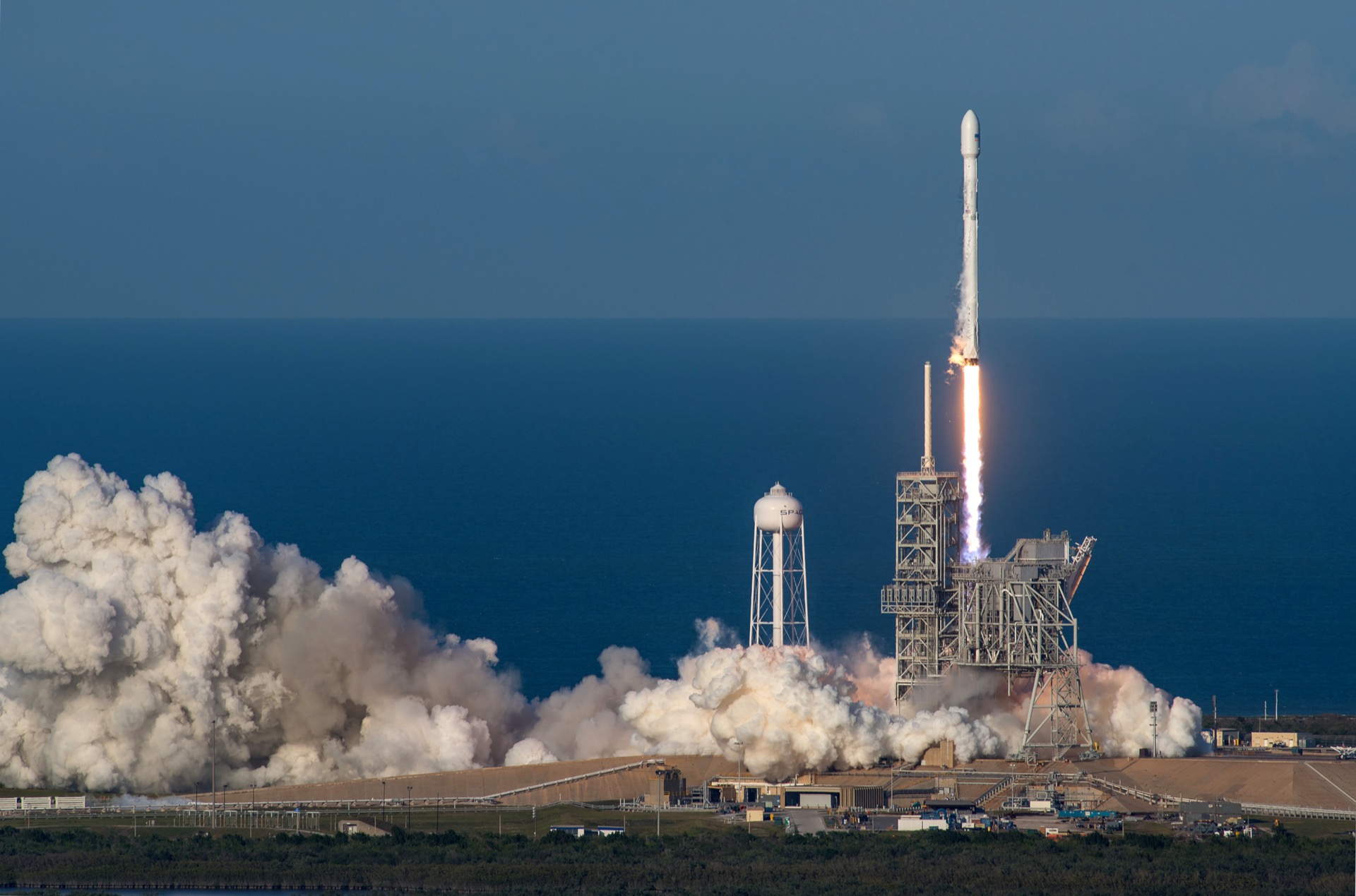A SpaceX Falcon 9 rocket lifts off from its coastal launch pad on a column of flame, with the SpaceX water tower and the Atlantic Ocean behind it
