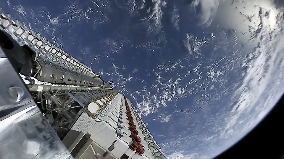 An onboard camera looks along a flat-packed stack of Starlink satellites attached to a Falcon 9 upper stage with Earth curving below