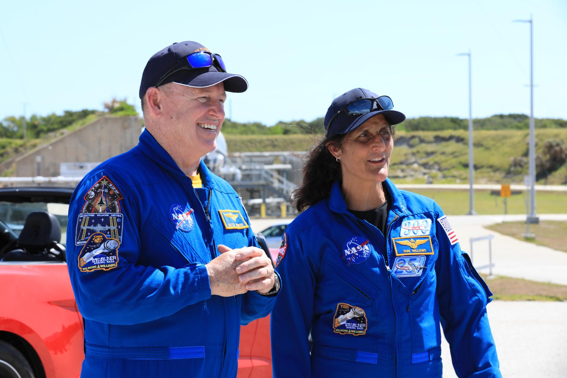 NASA astronauts Butch Wilmore and Suni Williams in blue flight suits with Starliner mission patches, smiling near the launch pad with a red convertible crew-transport car behind them