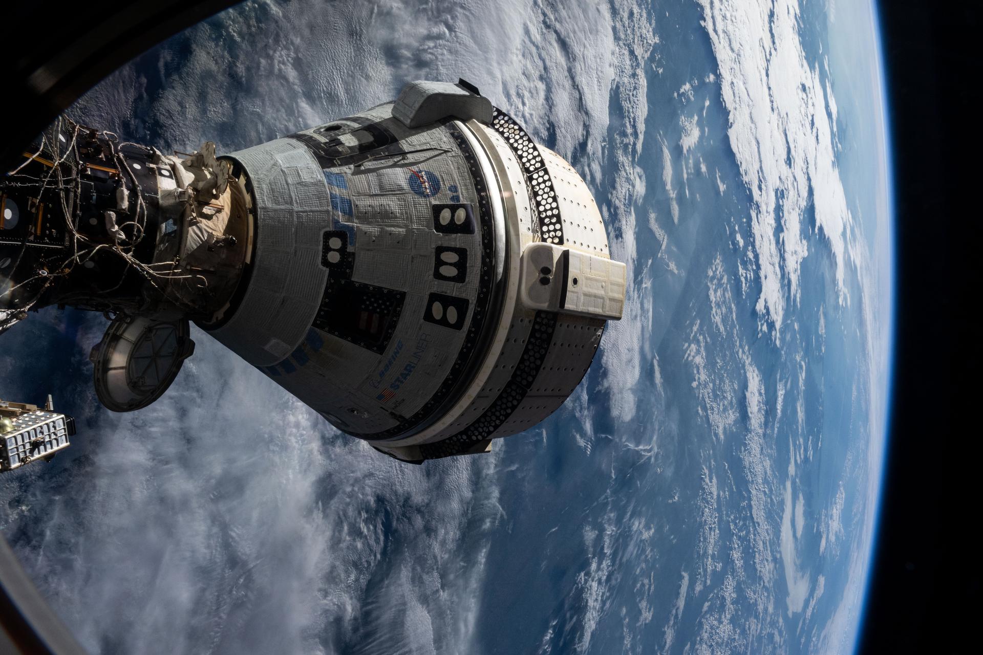 Close-up of Boeing's Starliner capsule docked to the International Space Station, the word STARLINER and a NASA logo visible on its silver-and-white hull, with Earth's blue-and-white surface curving below