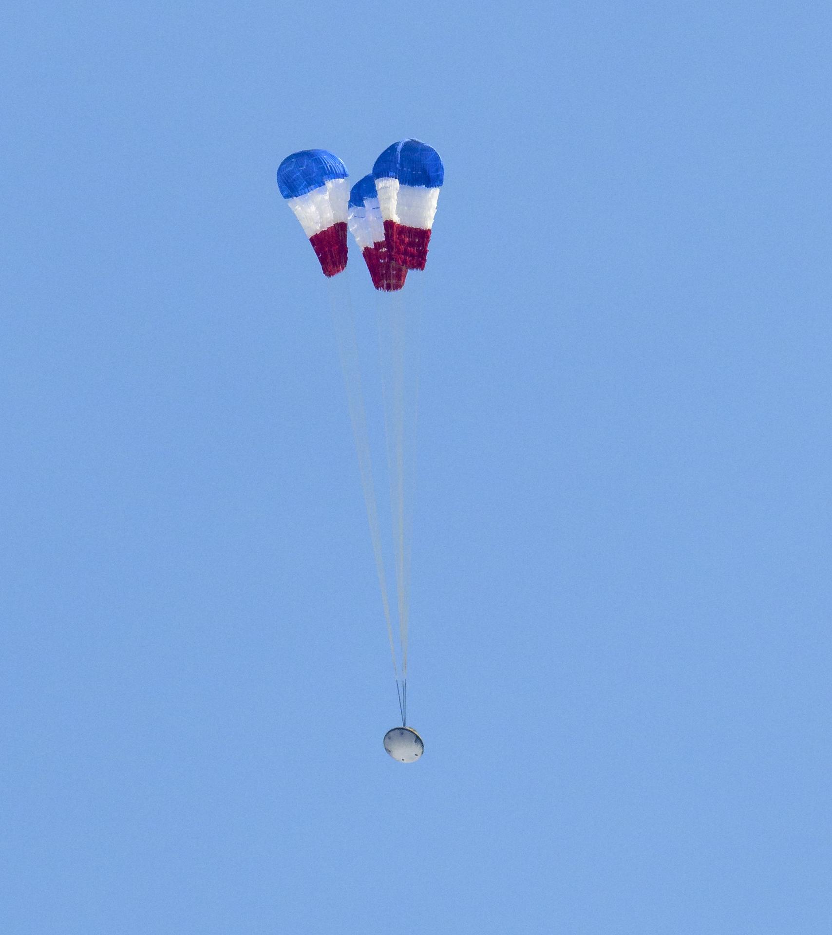 A Starliner capsule descending under three red-white-and-blue main parachutes against a clear blue sky
