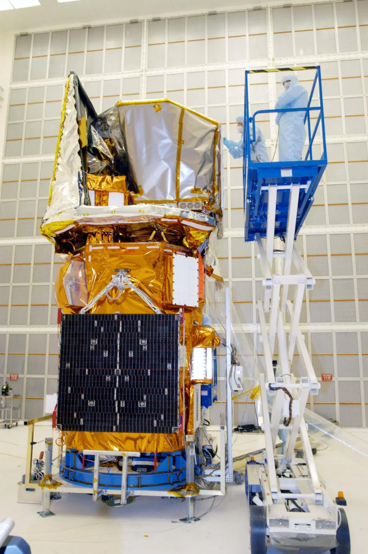 The Swift observatory standing in a clean high bay wrapped in gold and silver thermal blankets, with two technicians in cleanroom suits on a blue lift platform beside it