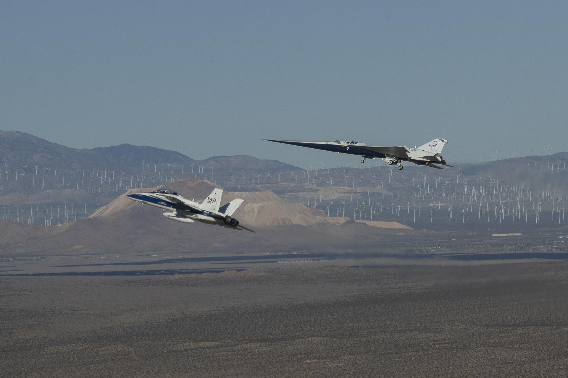 NASA's X-59 quiet supersonic aircraft with landing gear down flying above the Mojave Desert alongside a NASA F/A-18 escort aircraft, with wind turbines and mountains visible below, during its first flight on October 28, 2025