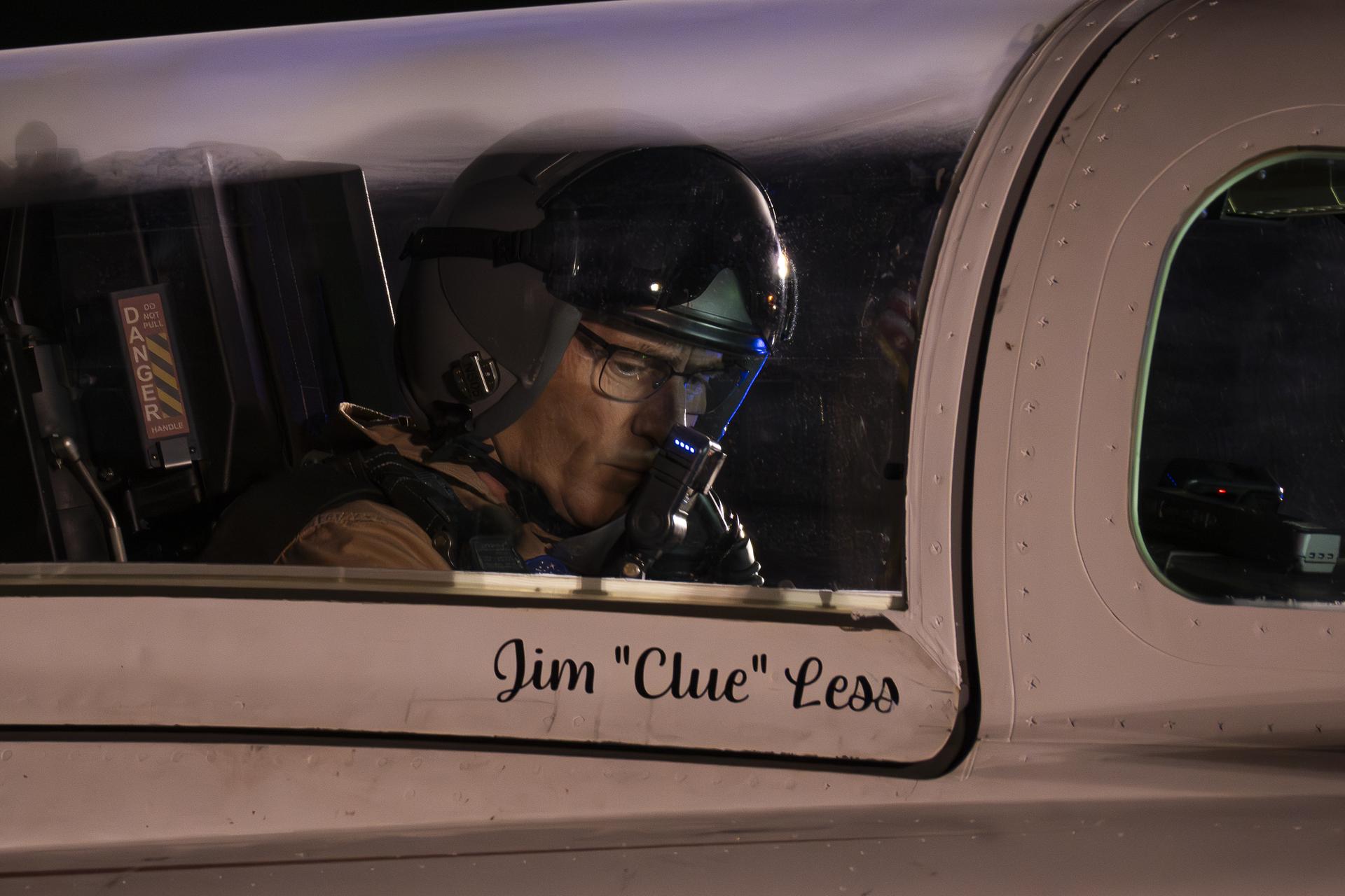 NASA test pilot Jim 'Clue' Less sits inside the X-59 cockpit in low light, wearing a flight helmet and glasses, with his name painted on the fuselage outside the closed canopy