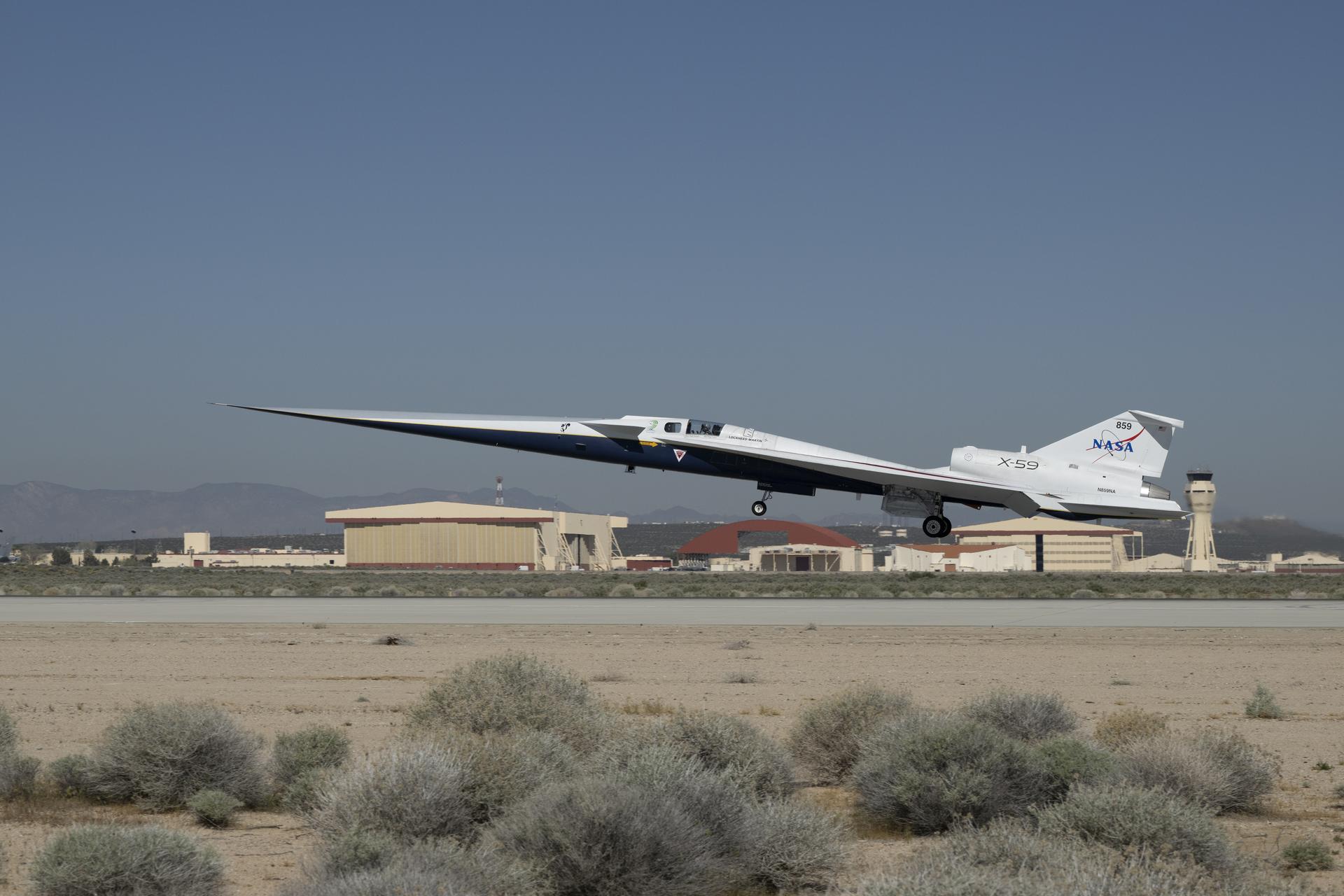 NASA's X-59 quiet supersonic aircraft lifting off from Edwards Air Force Base on March 20, 2026, showing the extremely long needle nose, delta wing, and NASA 859 tail markings, with desert scrub in the foreground and base hangars behind