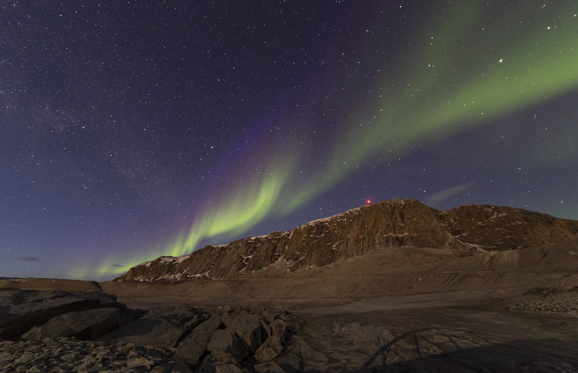Green and purple aurora over a rocky, snow-patched ridge in Greenland
