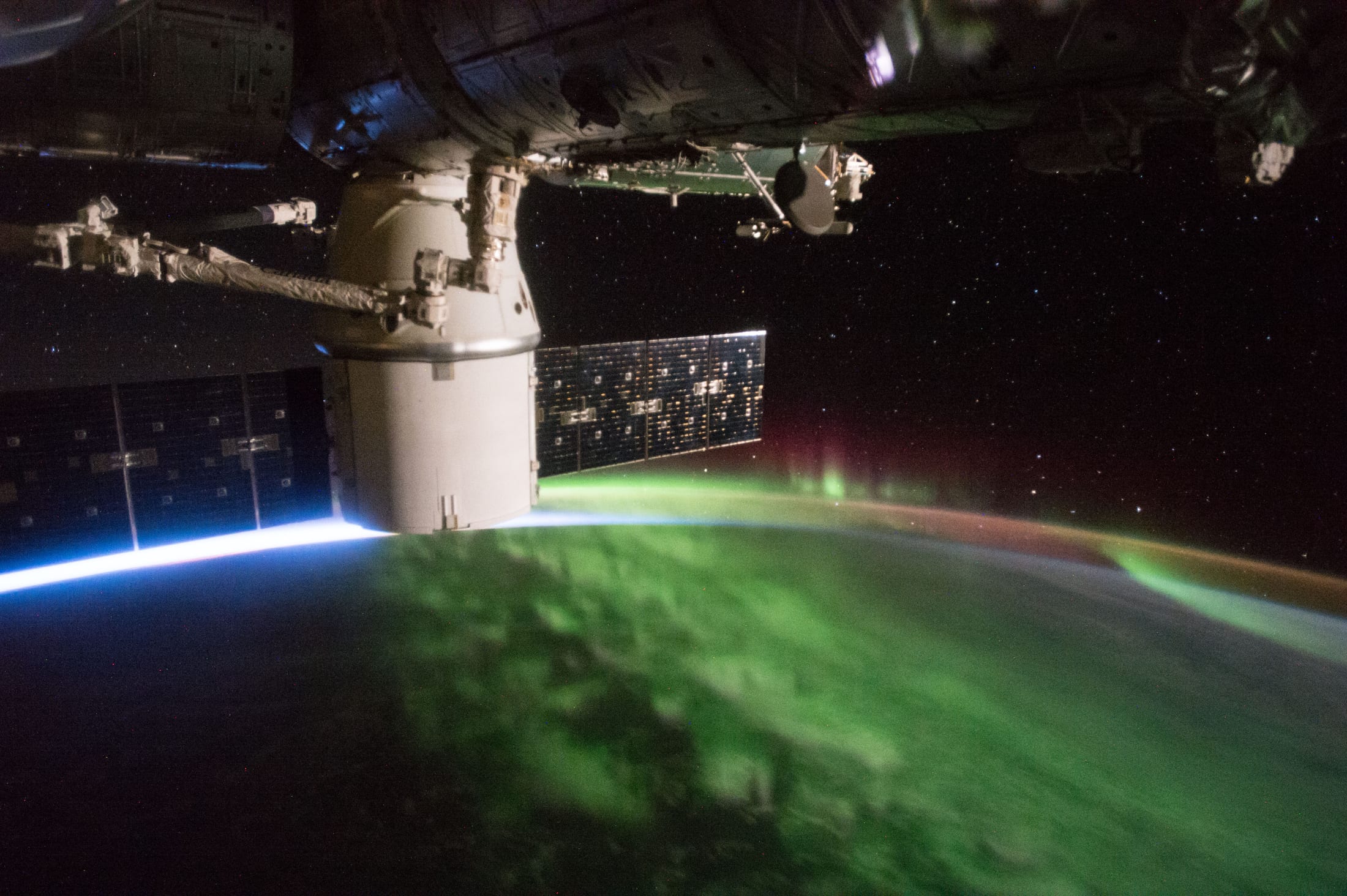 Green and red aurora on Earth's limb framed by a SpaceX Dragon capsule and ISS solar array