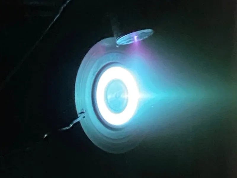 A Bellatrix Aerospace Hall-effect thruster fires in a vacuum chamber, emitting its characteristic blue-white plasma plume — the company's signature electric-propulsion technology.