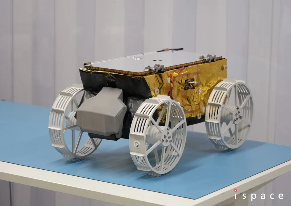 Side view of ispace's TENACIOUS lunar micro rover — gold-foil body and four white wheels — on a display stand at ispace-EUROPE in Luxembourg.