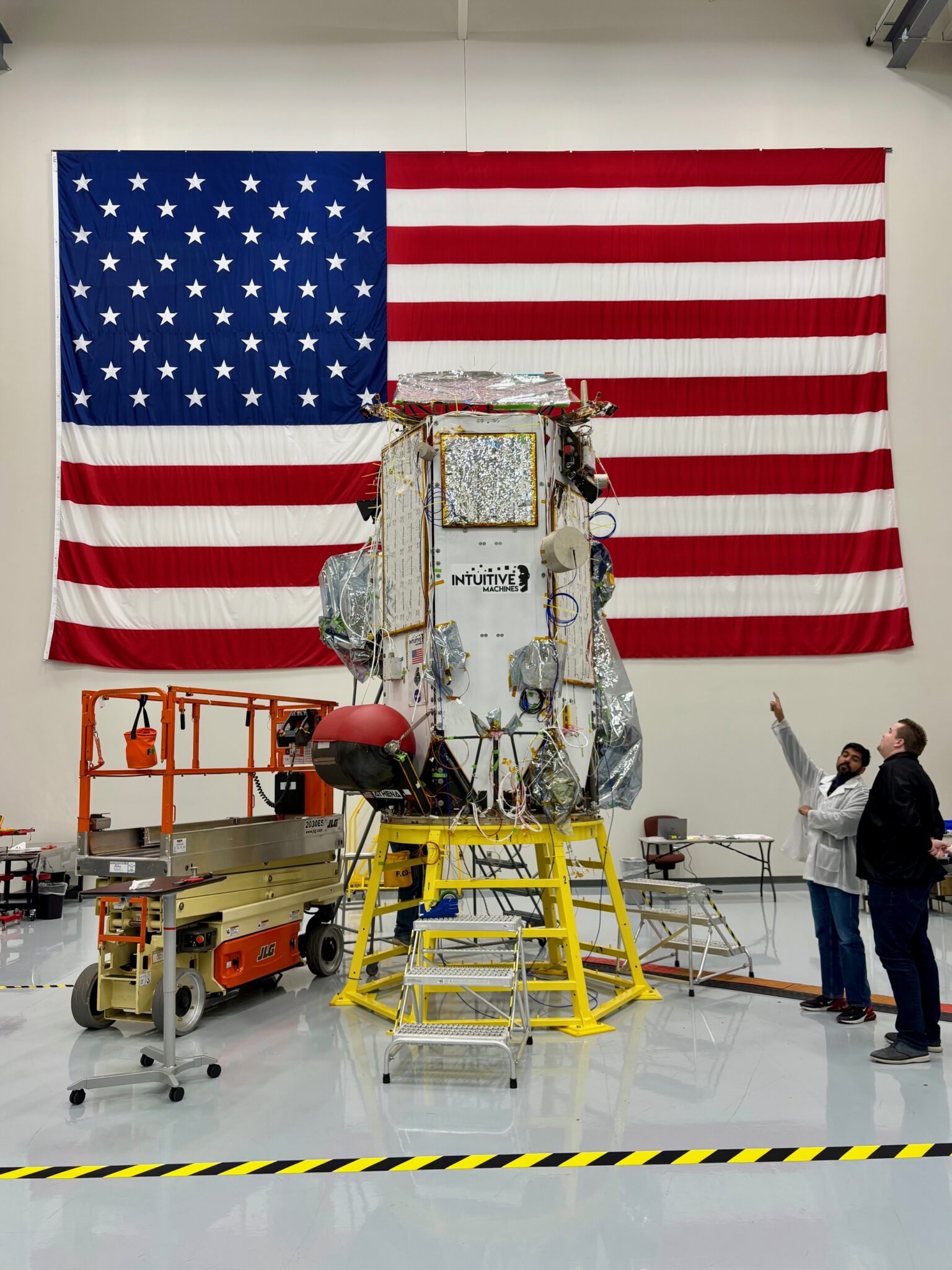 Odysseus Nova-C lander during final assembly at Intuitive Machines' Houston facility, in front of the American flag — the lander that returned the US to the Moon